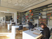 Blick in einen Lesesaal. Die Bücherregale stehen an der Wand, davor sieht man moderne Arbeitsplätze, an denen Menschen sitzen und lesen.