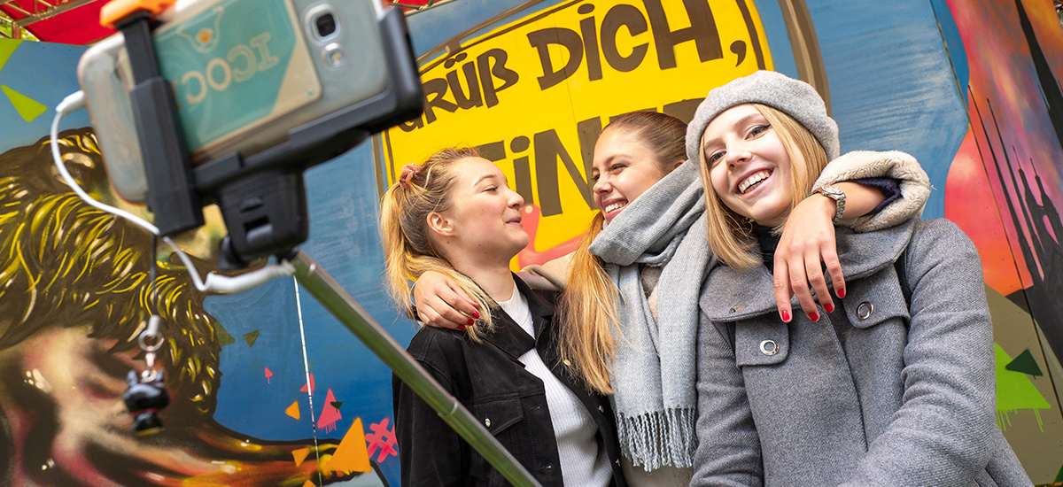 Drei junge Studierende machen mit einem Selfiestick ein Erinnerungsfoto vor der Selfiewand.