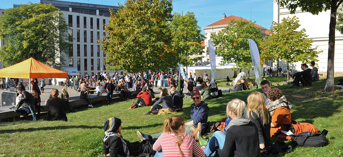 Studierende genießen auf einer Wiese die letzten warmen Sonnenstrahlen im Oktober. Dabei blicken sie auf das bunte Treiben vor den Infoständen.