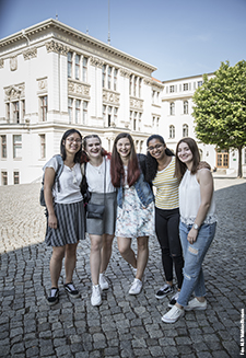 Auf dem Foto sieht man 5 junge Frauen, Arm in Arm auf einem Campus.