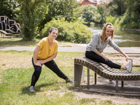 Das Bild zeigt zwei Studentinnen beim Stretching im Park an einer Bank.