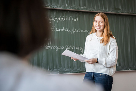 Auf dem Bild seht ihr, wie ich mit einem Hefter in der Hand vor der Tafel stehe. Man erkennt den Schriftzug "Differentialgleichungen" an der Tafel.