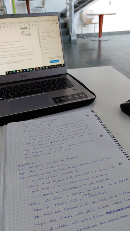 Auf dem Bild sieht man einen Laptop und einen karierten Block davor mit Aufgaben zur Vorlesung Zahlentheorie. Im Hintergrund kann man das Cafe Einstein am Heide-Campus erkennen.