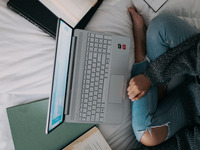 Zu sehen ist eine Frau, die auf dem Bett sitzt. Vor ihr liegen viele Bücher und ihr Laptop. Sie schreibt an einer Hausarbeit.