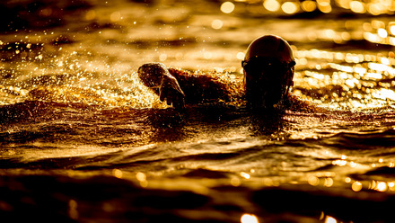 Das Bild zeigt eine Nahaufnahme eines Schwimmers im Wasser. Er hat eine Schwimmkappe auf und das Licht, das sich im Wasser bricht ist orangegelb.