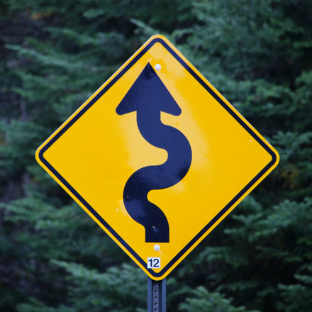 Zu sehen ist ein Straßenschild, auf dem ein krummer Pfeil eine ungerade verlaufende Strecke anzeigt.