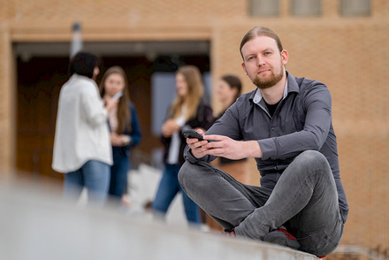 Zu sehen ist Dr. Busch mit einem Smartphone in der Hand. Im Hintergrund steht eine Gruppe von Jugendlichen.