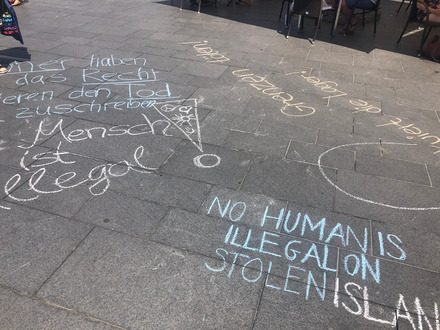 Auf dem Foto sieht man verschiedene Schriftzüge, die mit Kreide auf den Boden gemalt wurden. Da steht zum Beispiel: Kein Mensch ist illegal.