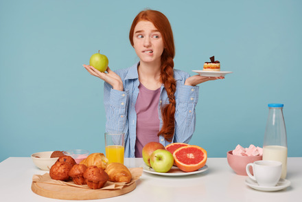 Auf dem Foto sieht man eine junge Frau, die einen Apfel und einen Kuchen in der Hand hält und verwirrt aussieht. Auf dem Tisch vor ihr liegen weitere Lebensmittel.