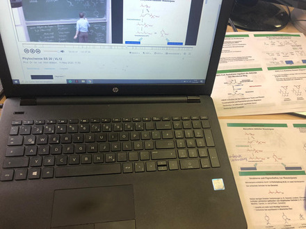 Auf dem Foto sieht man einen Laptop, auf dem eine online Vorlesung läuft und einige Seiten eines Skriptes.