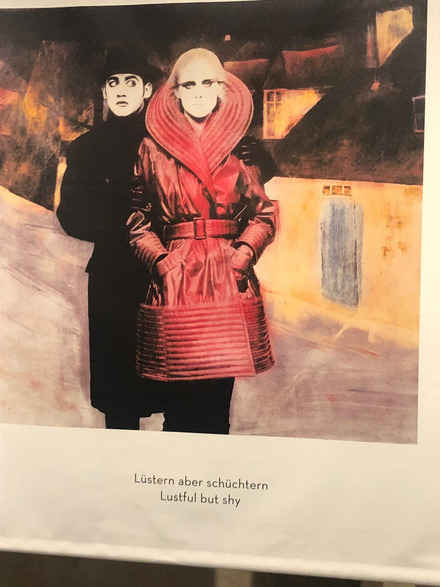 Auf dem Foto sieht man eine Frau in einem rotem Mantel die von einem Herren im schwarzen Mantel von hinten umarmt wird. Es ist eine künstlerische Fotografie von Karl Lagerfeld.