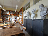 In der Mitte stehen große Tische zum Arbeiten. An den Wänden stehen hohe Bücherregale und einige Gelehrtenbüsten.