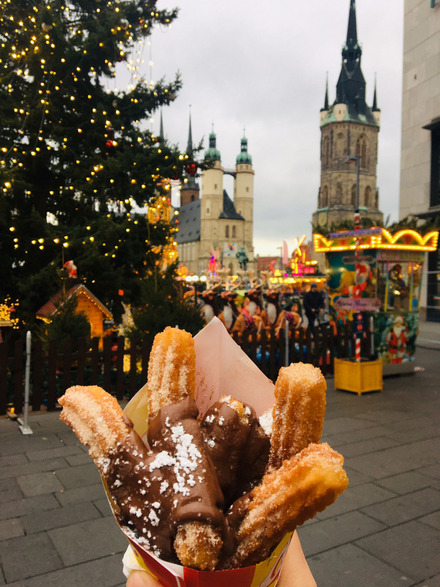 Zu sehen ist der Weihnachtsmarkt in Halle im Hintergrund und eine Hand, die eine Tüte voller Churros mit Nutella in der Hand hält.