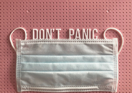 Zu sehen ist eine medizinische Gesichtsmaske vor rosa Hintergrund und vor dem Schriftzug "Don't Panic" (Keine Panik).