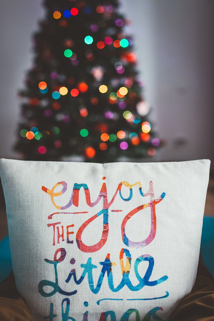 Zu sehen ist ein Kissen mit dem Aufdruck "Enjoy the little things" und im Hintergrund ein funkelnder Weihnachtsbaum.