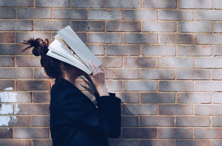Zu sehen ist eine Studentin, welche sich ein Buch vors Gesicht hält.